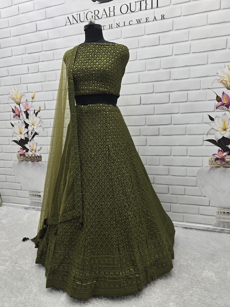 May include: An olive green ethnic outfit, including a crop top and a long skirt, displayed on a mannequin. The outfit features intricate patterns and a matching sheer scarf. The background is a white brick wall. The text "ANUGRAH OUTFIT" is visible at the top.