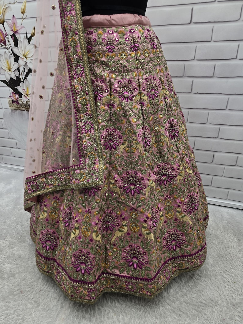 May include: A pink and gold embroidered lehenga skirt with intricate floral and bird patterns. The skirt features a detailed border and is paired with a matching dupatta. The fabric has a shimmering effect, and the design is rich with embellishments.