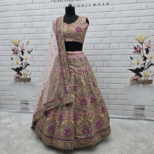 May include: A pink and gold embroidered lehenga choli set on a mannequin. The set includes a skirt, a cropped top, and a sheer dupatta. The outfit features intricate floral embroidery and embellishments. The text "ANUGRAH OUTFIT ETHNICWEAR" is visible in the background.