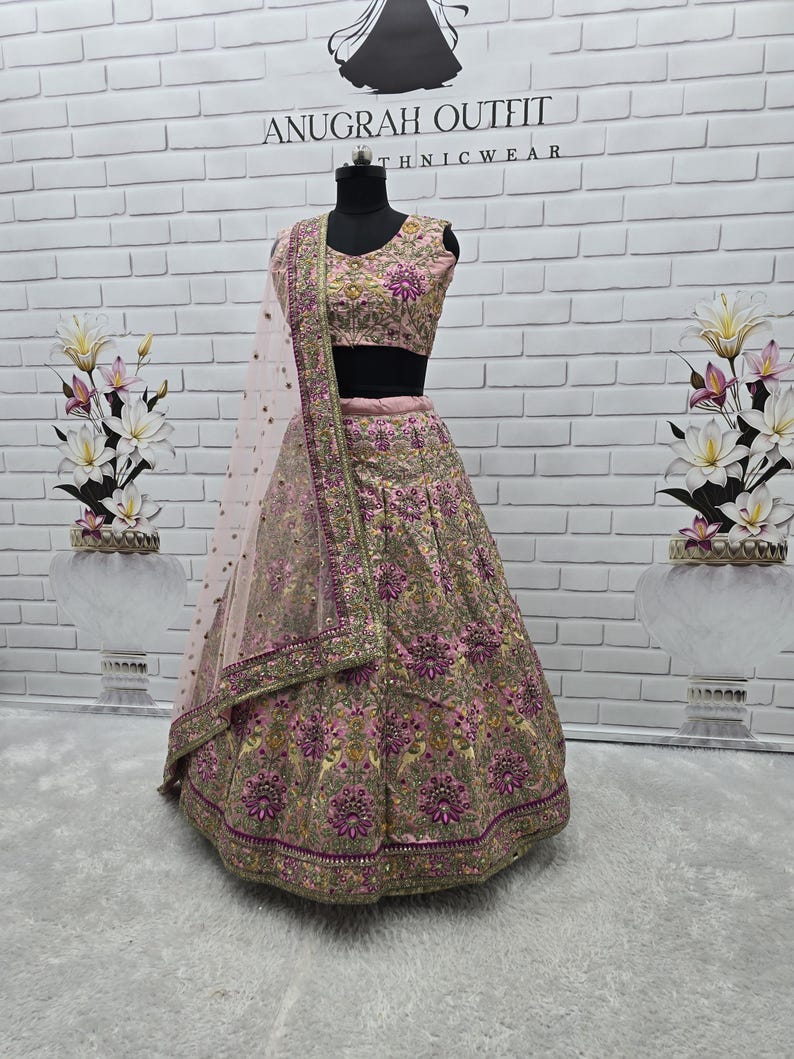 May include: A pink and gold embroidered lehenga choli set on a mannequin. The set includes a skirt, a cropped top, and a sheer dupatta. The outfit features intricate floral embroidery and embellishments. The text "ANUGRAH OUTFIT ETHNICWEAR" is visible in the background.