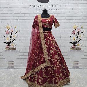 May include: A burgundy ethnic outfit, including a top and skirt, displayed on a mannequin. The outfit features intricate gold embroidery and a matching sheer scarf. The background is a white brick wall with the text "ANUGRAH OUTFIT ETHNIC WEAR".