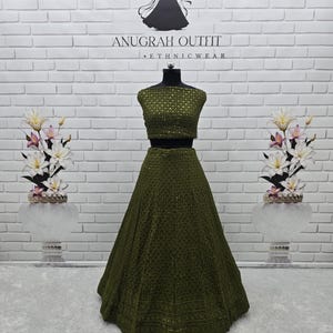 May include: An olive green two-piece ethnic wear outfit on a mannequin. The top is a fitted crop top, and the skirt is a full, flowing A-line. The outfit is embellished with intricate patterns. The background features a white brick wall and floral arrangements.