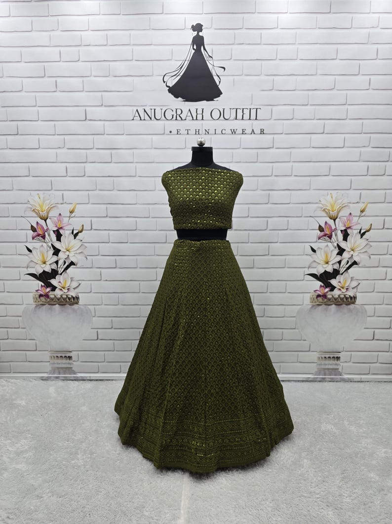 May include: An olive green two-piece ethnic wear outfit on a mannequin. The top is a fitted crop top, and the skirt is a full, flowing A-line. The outfit is embellished with intricate patterns. The background features a white brick wall and floral arrangements.