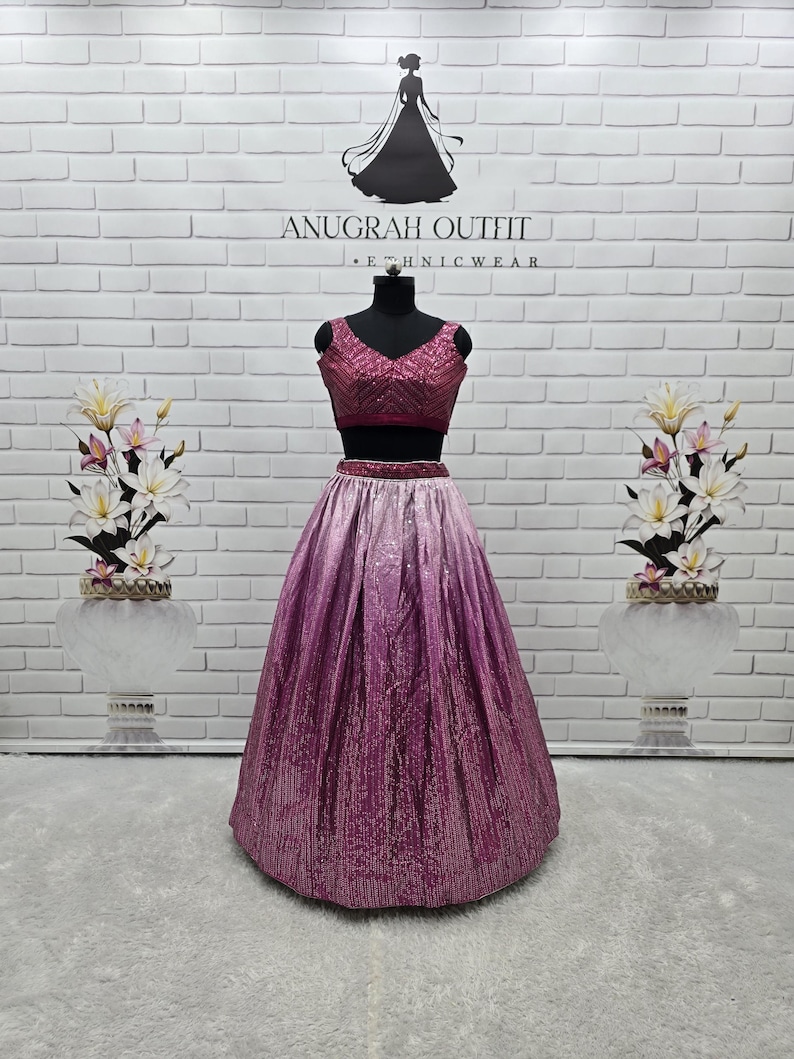 May include: A two-piece formal outfit on a mannequin. The top is a deep pink, sequined crop top with a V-neck. The skirt is a long, ombre design, transitioning from light pink to deep pink. The outfit is displayed in front of a white brick wall.