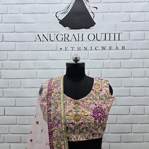 May include: A light pink ethnic outfit with intricate embroidery and embellishments. The top features floral and bird motifs, with a matching dupatta. The outfit is displayed on a black mannequin against a white brick wall. The brand name "ANUGRAH OUTFIT" is visible.