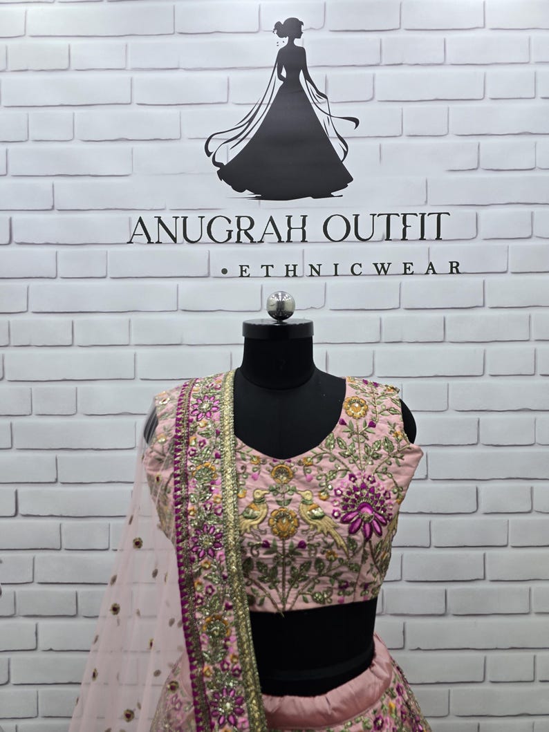 May include: A light pink ethnic outfit with intricate embroidery and embellishments. The top features floral and bird motifs, with a matching dupatta. The outfit is displayed on a black mannequin against a white brick wall. The brand name "ANUGRAH OUTFIT" is visible.