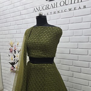 May include: An olive green ethnic outfit, displayed on a mannequin. The outfit includes a crop top, skirt, and a sheer, dotted shawl. The top and skirt feature a detailed, textured pattern. The text "ANUGRAH OUTFIT ETHNICWEAR" is visible on the wall.