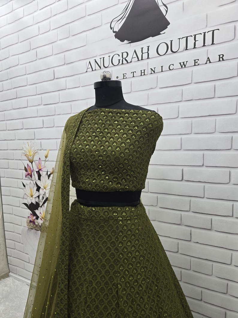 May include: An olive green ethnic outfit, displayed on a mannequin. The outfit includes a crop top, skirt, and a sheer, dotted shawl. The top and skirt feature a detailed, textured pattern. The text "ANUGRAH OUTFIT ETHNICWEAR" is visible on the wall.