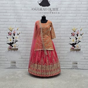 May include: An orange and pink ethnic outfit with intricate gold embroidery. The outfit includes a long-sleeved top and a layered skirt. A sheer pink scarf is draped over the shoulder. The text "ANUGRAH OUTFIT - ETHNICWEAR" is visible.