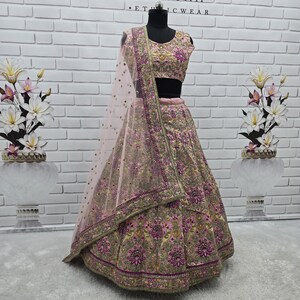 May include: A pink and gold embroidered lehenga choli set on a mannequin. The set includes a sleeveless top, a long skirt, and a sheer dupatta. The outfit is adorned with intricate floral and geometric patterns. The background features a white brick wall and the text "ANUGRAH OUTFIT".