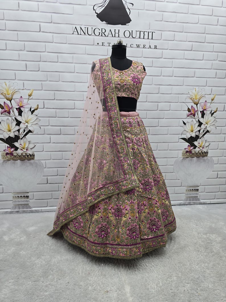 May include: A pink and gold embroidered lehenga choli set on a mannequin. The set includes a sleeveless top, a long skirt, and a sheer dupatta. The outfit is adorned with intricate floral and geometric patterns. The background features a white brick wall and the text "ANUGRAH OUTFIT".