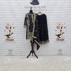 May include: Black ethnic outfit with gold embroidery, displayed on a mannequin. The outfit includes a long-sleeved top and a skirt. The background features a white brick wall and the text "ANUGRAH OUTFIT THNICWEAR".