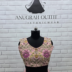 May include: A light pink embroidered ethnic outfit, including a top and skirt, displayed on a black mannequin. The top features a V-neck and intricate floral and bird embroidery with gold and purple accents. The background is a white brick wall with the text "ANUGRAH OUTFIT . ETHNICWEAR".