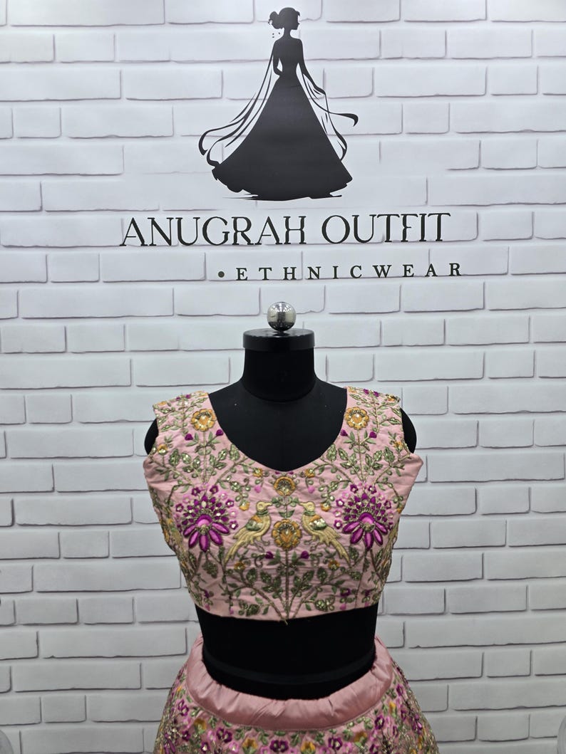 May include: A light pink embroidered ethnic outfit, including a top and skirt, displayed on a black mannequin. The top features a V-neck and intricate floral and bird embroidery with gold and purple accents. The background is a white brick wall with the text "ANUGRAH OUTFIT . ETHNICWEAR".