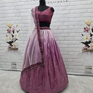 May include: A formal, two-piece outfit in shades of pink and purple, displayed on a mannequin. The top is a deep magenta, and the skirt transitions from light pink to a darker purple. A sheer, light pink scarf with a magenta border completes the ensemble. The outfit is labeled "ANUGRAH OUTFIT".
