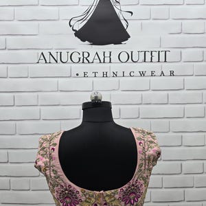 May include: A light pink embroidered ethnic wear top with floral and leaf patterns in shades of purple, green, and gold. The top is displayed on a black mannequin against a white brick wall. The text "ANUGRAH OUTFIT - ETHNIC WEAR" is visible.