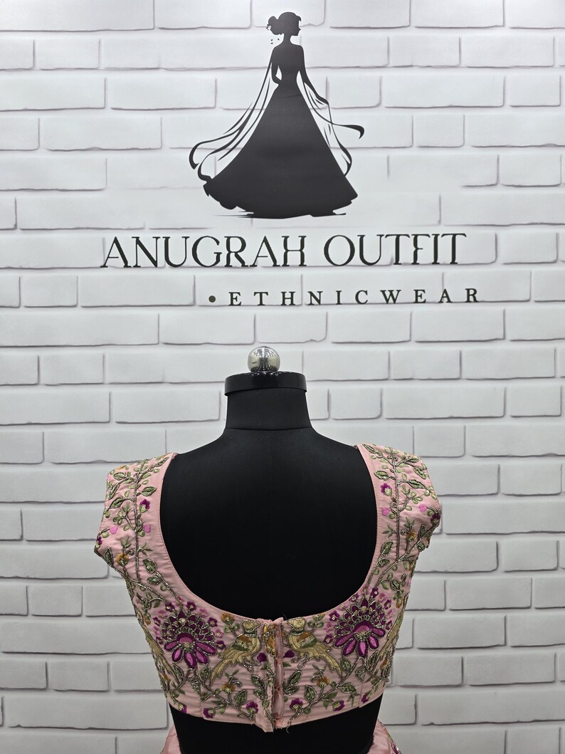 May include: A light pink embroidered ethnic wear top with floral and leaf patterns in shades of purple, green, and gold. The top is displayed on a black mannequin against a white brick wall. The text "ANUGRAH OUTFIT - ETHNIC WEAR" is visible.