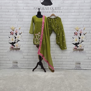 May include: An olive green ethnic wear outfit, comprising a tunic and trousers, displayed on a mannequin. The tunic has floral embroidery and is paired with a pink and white striped scarf. The outfit is set against a white brick wall.