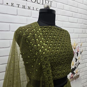 May include: An olive green ethnic outfit displayed on a mannequin. The top features intricate sequin detailing and a matching sheer scarf. The outfit is set against a white brick wall with the text "JGRAH OUTFIT ETHNIC".