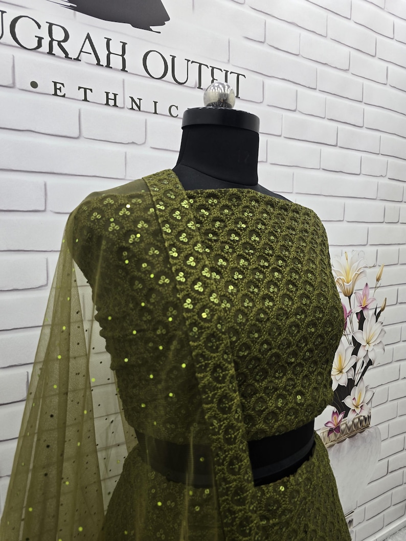 May include: An olive green ethnic outfit displayed on a mannequin. The top features intricate sequin detailing and a matching sheer scarf. The outfit is set against a white brick wall with the text "JGRAH OUTFIT ETHNIC".