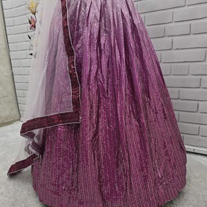 May include: A magenta and purple ombre skirt with a matching embellished waistband. The skirt is covered in small, reflective sequins, creating a shimmering effect. A sheer, white dupatta with a magenta border is draped over the skirt.
