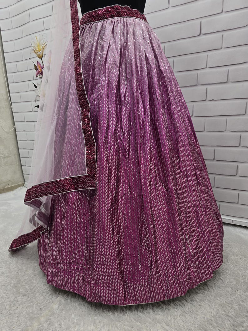 May include: A magenta and purple ombre skirt with a matching embellished waistband. The skirt is covered in small, reflective sequins, creating a shimmering effect. A sheer, white dupatta with a magenta border is draped over the skirt.