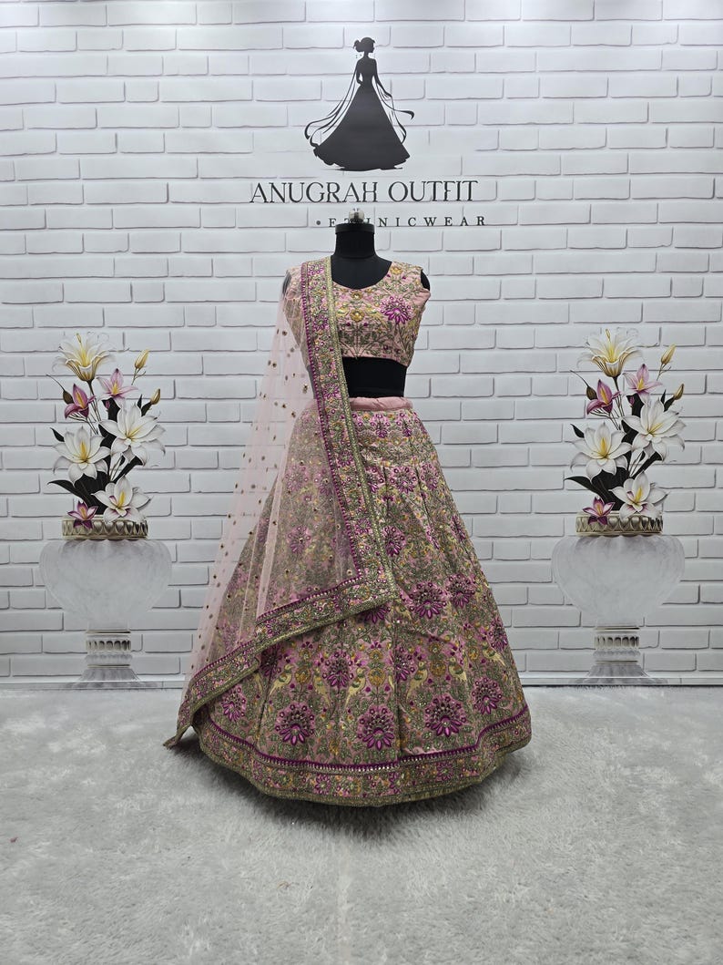 May include: A pink and gold embroidered lehenga choli set. The set includes a top, skirt, and a sheer dupatta. The outfit is adorned with intricate floral patterns and embellishments. The background features the text "ANUGRAH OUTFIT" and a silhouette of a woman in a gown.