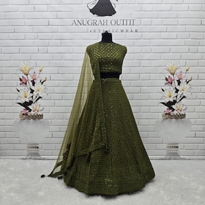 May include: Olive green ethnic wear outfit, including a top, skirt, and sheer scarf. The outfit is embellished with intricate patterns and sequins. The text "ANUGRAH OUTFIT - ETHNIC WEAR" is visible on the background.