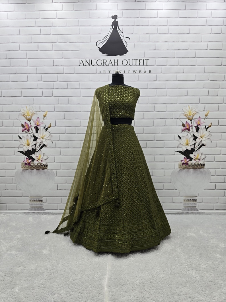 May include: Olive green ethnic wear outfit, including a top, skirt, and sheer scarf. The outfit is embellished with intricate patterns and sequins. The text "ANUGRAH OUTFIT - ETHNIC WEAR" is visible on the background.