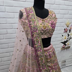 May include: A light pink embroidered ethnic outfit, including a top and skirt, displayed on a mannequin. The outfit features intricate floral and bird embroidery in shades of purple, gold, and green. A sheer, light pink scarf is draped over the shoulder.