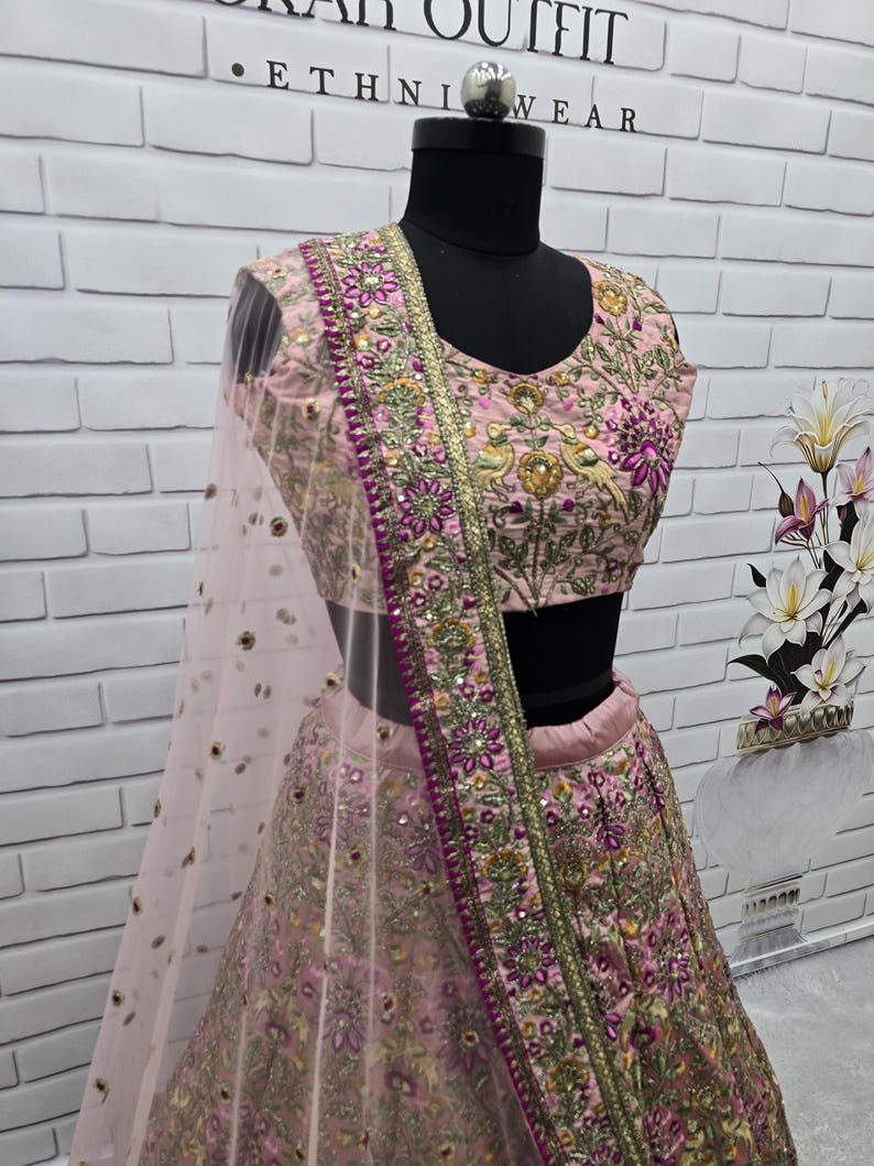 May include: A light pink embroidered ethnic outfit, including a top and skirt, displayed on a mannequin. The outfit features intricate floral and bird embroidery in shades of purple, gold, and green. A sheer, light pink scarf is draped over the shoulder.