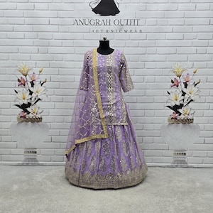 Op de afbeelding: Een lavendelkleurige etnische outfit met gouden versieringen, gepresenteerd op een mannequin. De outfit bestaat uit een top, rok en een doorschijnende sjaal. De tekst "ANUGRAH OUTFIT ETHNICWEAR" is zichtbaar bovenaan de afbeelding.