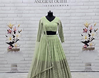 May include: A pale green ethnic outfit, comprising a long-sleeved top and a flowing skirt, presented on a mannequin. The outfit is adorned with detailed embellishments and a matching sheer scarf. The backdrop features a white brick wall and floral arrangements. The text "ANUGRAH OUTFIT - ETHNICWEAR" is displayed.