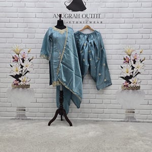 May include: A dusty blue three-piece outfit consisting of a long-sleeved tunic with gold embroidery around the neckline and hem, a matching draped dupatta, and pleated trousers. The set is displayed on a mannequin and a wooden hanger.