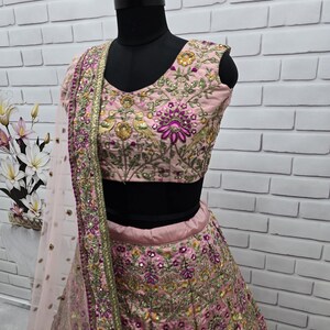 May include: A light pink ethnic wear ensemble, featuring a heavily embroidered top and skirt. The outfit is adorned with intricate floral patterns in shades of purple, gold, and green. A matching sheer dupatta with a gold border completes the look. The mannequin is set against a white brick wall.