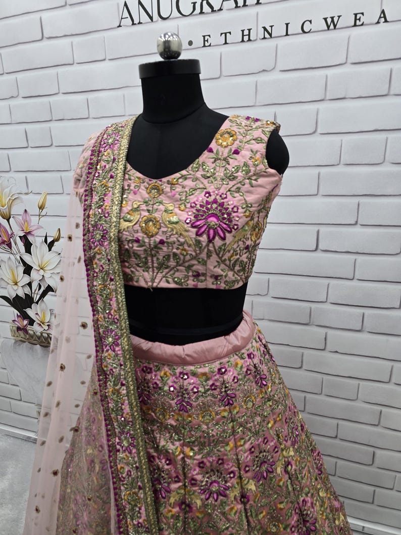 May include: A light pink ethnic wear ensemble, featuring a heavily embroidered top and skirt. The outfit is adorned with intricate floral patterns in shades of purple, gold, and green. A matching sheer dupatta with a gold border completes the look. The mannequin is set against a white brick wall.