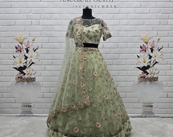 May include: A light green lehenga choli set featuring a sheer dupatta and blouse with intricate floral embroidery in pink and gold tones. The skirt has a wide hemline with matching floral patterns. The text ANUGRAH OUTFIT is visible.