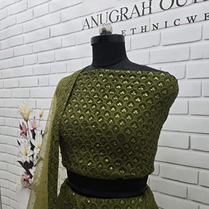May include: An olive green ethnic wear ensemble, displayed on a mannequin. The outfit features intricate sequin detailing and a matching sheer dupatta. The top has a round neckline and a cropped silhouette. The brand name "ANUGRAH OUTWEAR ETHNIC WE" is visible in the background.