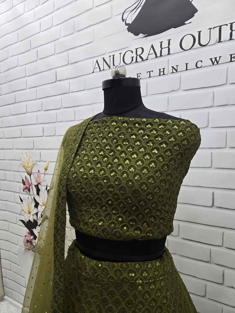 May include: An olive green ethnic wear ensemble, displayed on a mannequin. The outfit features intricate sequin detailing and a matching sheer dupatta. The top has a round neckline and a cropped silhouette. The brand name "ANUGRAH OUTWEAR ETHNIC WE" is visible in the background.