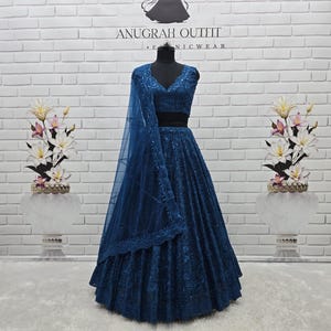 May include: A dark teal lehenga choli ensemble on a mannequin. The outfit features intricate embroidery and a matching dupatta. The background includes a white brick wall with the text "ANUGRAH OUTFIT" and floral arrangements.