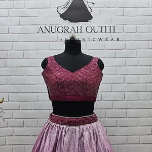 May include: A two-piece formal outfit with a burgundy top and a light purple skirt. The top has a V-neck and intricate embroidery. The skirt is a flowing design with a sequined pattern. The outfit is displayed on a black mannequin. The brand name "ANUGRAH OUTFIT" is visible.