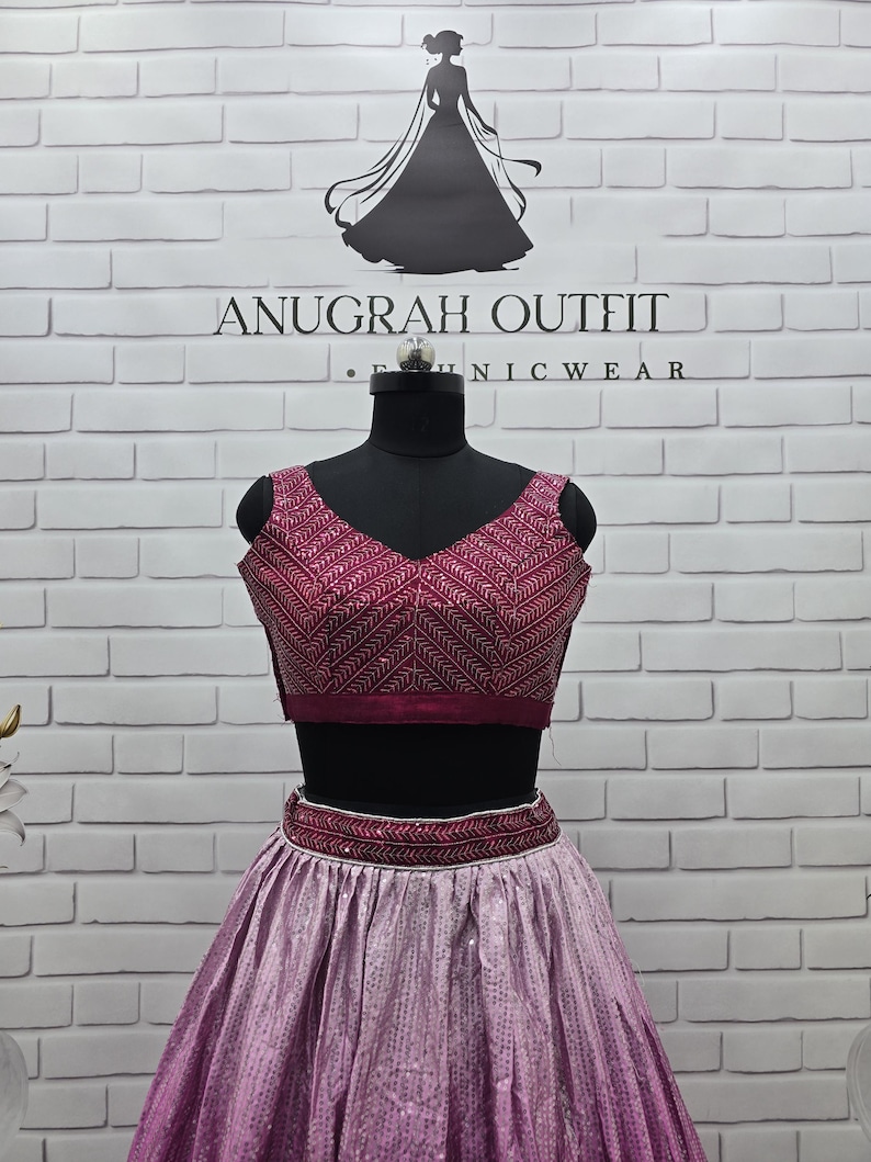 May include: A two-piece formal outfit with a burgundy top and a light purple skirt. The top has a V-neck and intricate embroidery. The skirt is a flowing design with a sequined pattern. The outfit is displayed on a black mannequin. The brand name "ANUGRAH OUTFIT" is visible.