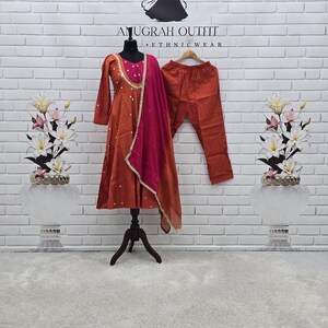 Peut inclure: Une robe à manches longues de couleur rouille avec une jupe évasée, ornée de décorations dorées et d'une dupatta magenta vif drapée sur l'épaule. L'ensemble comprend un pantalon assorti de couleur rouille présenté sur un mannequin.
