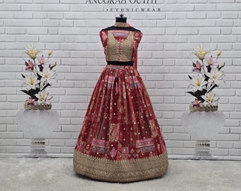 May include: A red and gold ethnic outfit displayed on a mannequin. The outfit features a detailed bodice and a full skirt with gold trim. The background includes the text "ANUGRAH OUTFIT - ETHNICWEAR" and decorative floral arrangements.