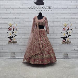 May include: A dusty rose-coloured ethnic outfit, featuring a long-sleeved top and a full skirt, adorned with intricate gold embroidery. A matching sheer scarf drapes over the shoulders. The outfit is displayed on a mannequin against a white brick wall. The text "ANUGRAH OUTFIT ETHNICWEAR" is visible.