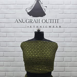 May include: An olive green ethnic outfit displayed on a mannequin. The outfit features a detailed, sequined pattern. The background is a white brick wall with the text "ANUGRAH OUTFIT - ETHNIC WEAR" and a silhouette of a woman in a dress.