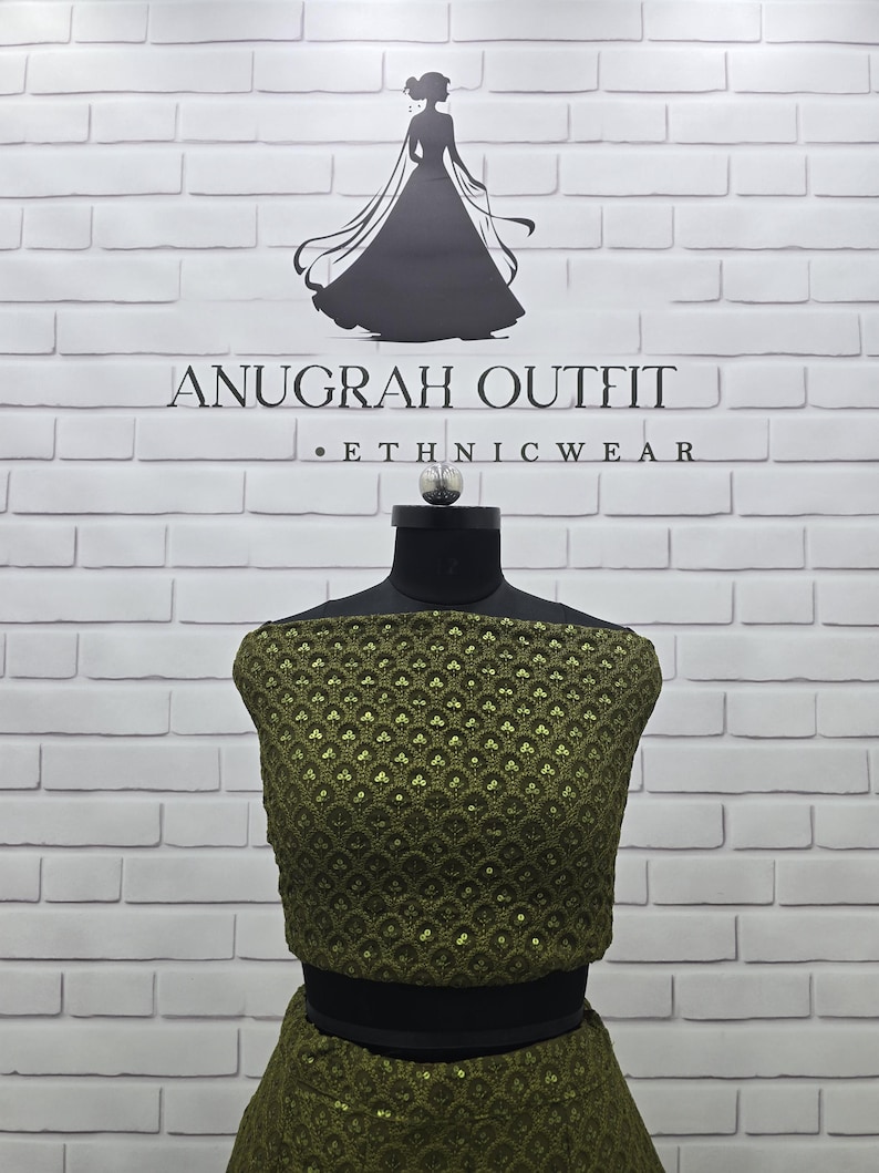 May include: An olive green ethnic outfit displayed on a mannequin. The outfit features a detailed, sequined pattern. The background is a white brick wall with the text "ANUGRAH OUTFIT - ETHNIC WEAR" and a silhouette of a woman in a dress.