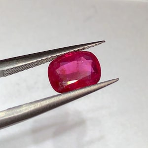 May include: A vibrant, cushion-cut ruby gemstone held by tweezers. The ruby displays a rich, deep red hue with a polished surface. The tweezers are silver-colored, contrasting with the gemstone's color. The background is a soft, neutral tone.