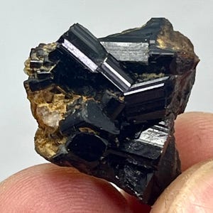 Op de afbeelding: Een close-up van een zwarte toermalijn kristalcluster met een glanzend, reflecterend oppervlak. Het kristal is geplaatst tegen een ruwe, bruinachtige matrix, die de natuurlijke vorm laat zien. Het donkere, ondoorzichtige mineraal heeft een opvallende uitstraling.