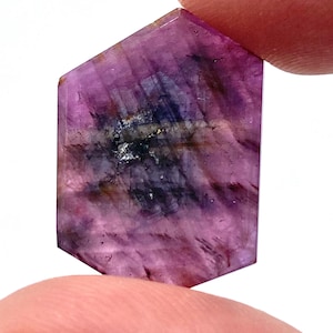 May include: A hexagonal-cut gemstone featuring a translucent magenta and deep purple color palette with visible internal inclusions, dark mineral streaks, and a polished surface that catches light across its flat, geometric faceted faces.